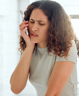 Woman in gray shirt in bathroom holding toothbrush and touching jaw in pain