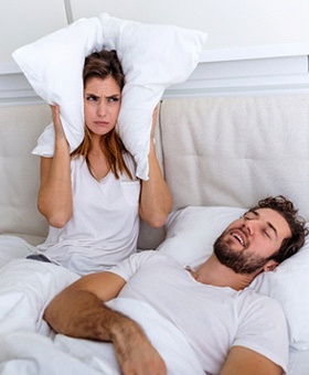 A man asleep in bed with his mouth open and snoring while the woman pushes a pillow against her ears to block out the noise